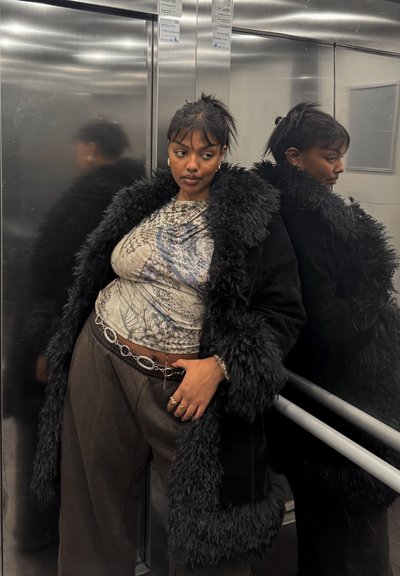 Abrigo de piel sintética negra con cuello texturizado, top gris estampado y pantalones oscuros holgados. Cinturón metálico de cadena con eslabones ovalados. Fondo de ascensor.