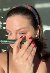 Mascara ad alto impatto Clinique in un tubetto verde, tenuto vicino al viso. La modella ha unghie rosse e una fascia per capelli a quadri; una luce solare soffusa crea ombre.