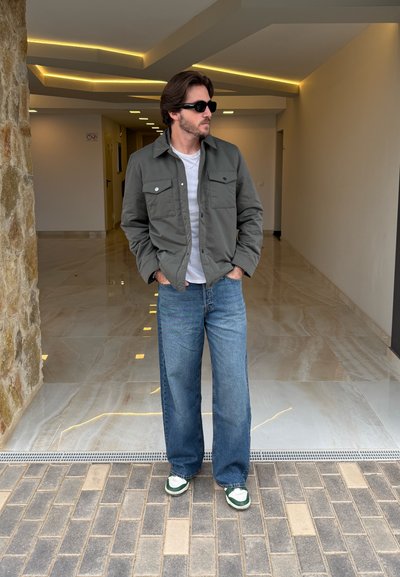 Chaqueta gris con dos bolsillos en el pecho, camiseta blanca, jeans azules de pierna ancha y zapatillas verdes. De pie en un entorno interior bien iluminado.