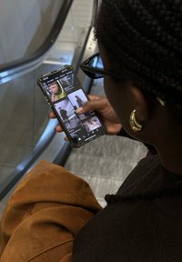Une personne tenant un smartphone avec une coque de protection, portant des lunettes à grosses montures, des boucles d'oreilles en or, et des vêtements en tissu brun texturé, sur un escalator.