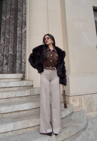 Chaqueta de piel sintética negra, camisa marrón de botones con bolsillo, pantalones de pierna ancha en un beige claro y zapatos oscuros de punta. Bolsa marrón con cadena dorada.