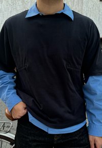 Top nero a maniche lunghe con una camicia celeste a collo visibile sotto, caratterizzato da due tasche frontali e una vestibilità morbida. Jeans in denim scuro indossati.