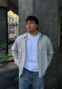 Codzienny strój z jasną wzorzystą koszulą i białym t-shirtem. Niebieskie dżinsy i teksturowane tło z granitu. Zielona barierka na pierwszym planie.