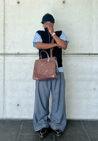 Bolso tote brillante en color marrón con un logo en relieve, combinado con pantalones anchos de rayas grises y una camiseta negra con cremallera y mangas azules.