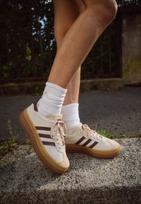 Sneaker in suede beige e tessuto con strisce marroni, dotati di una suola in gomma spessa. Calzini bianchi a coste visibili sopra la caviglia.