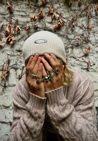 Beanie lavorato a maglia crema con logo, cardigan beige a maglia cable, molteplici anelli d'argento sulle mani, parete texturizzata con viti e foglie secche sullo sfondo.
