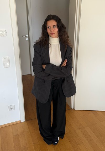 Blazer oversized a rayas negras sobre un cuello de tortuga blanco, combinado con pantalones holgados negros y zapatillas oscuras, de pie en una puerta.
