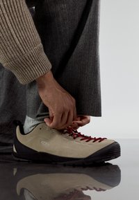 Beige suede shoe with red laces and black rubber sole. Close-up of a hand adjusting the laces, with textured grey trousers and patterned socks visible.