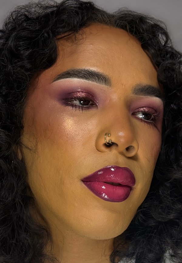 Gros plan d'une personne aux cheveux bouclés portant un rouge à lèvres brillant rose foncé, un fard à paupières violet et un anneau de nez en or dans la narine gauche.