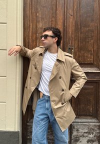 Trench coat beige con bottoni doppi, t-shirt bianca e jeans azzurri chiari davanti a una porta di legno. Include occhiali da sole e una posa casual.