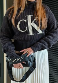 Personne portant un pull noir côtelé Calvin Klein et une jupe plissée blanche tenant un sac noir Tommy avec des bagues dorées aux doigts.