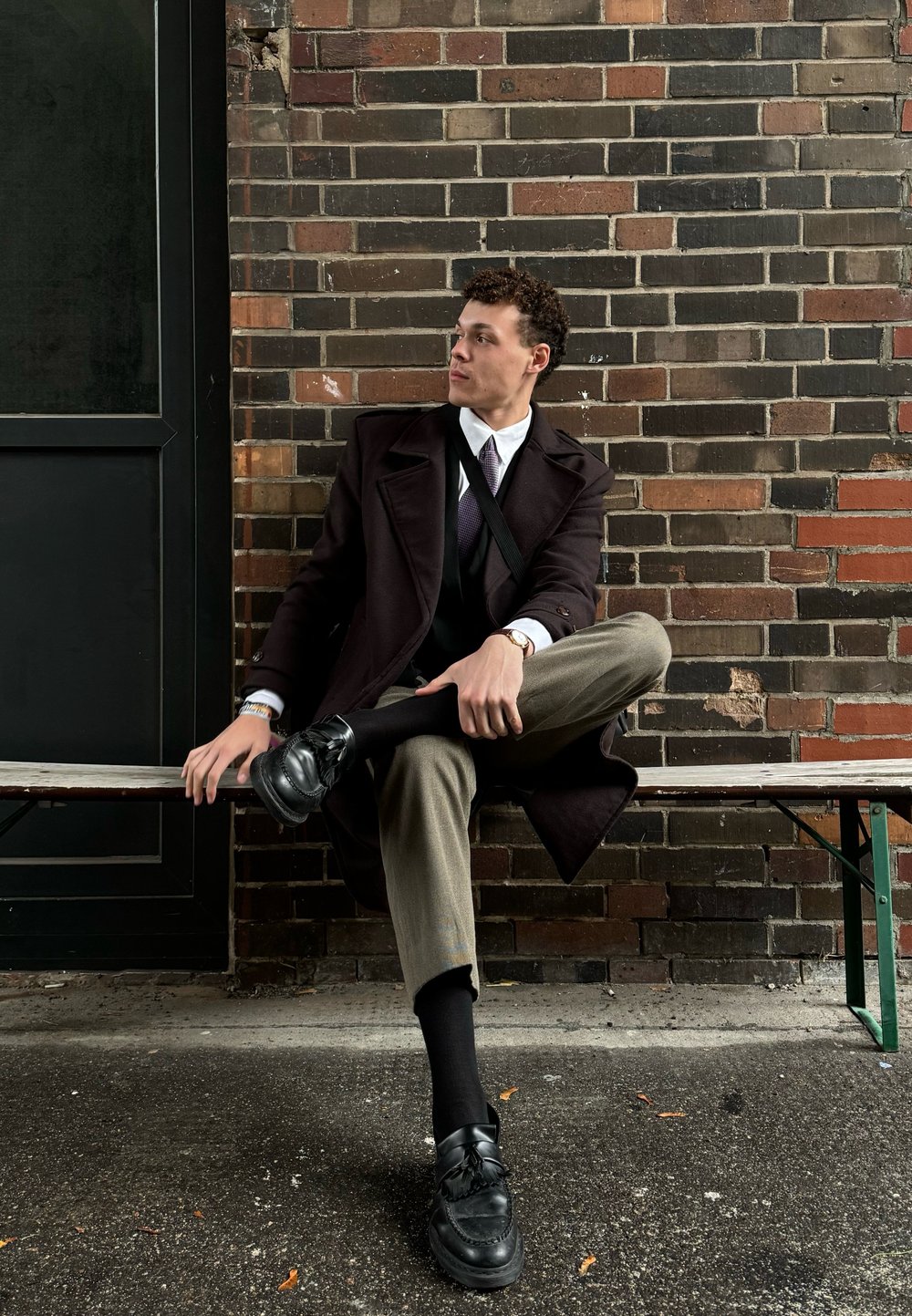 Μαύρο παλτό με δομημένους ώμους πάνω από σκούρο γιλέκο και λευκό πουκάμισο. Ελιά παντελόνι και μαύρα loafers. Φόντο από τοίχ�ο τούβλου.