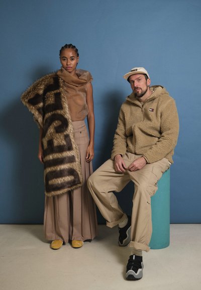 Dos modelos posan juntas. Una lleva una blusa texturizada marrón y una falda larga beige, drapeada con una bufanda de piel sintética con texto negro. La otra lleva un forro polar beis.