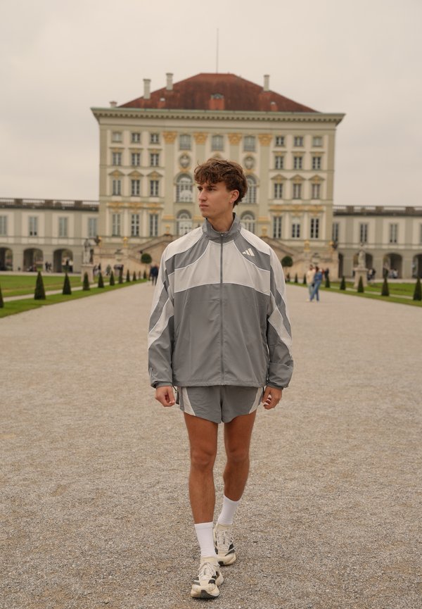 Un jeune homme en survêtement gris et blanc traverse la cour d’un palais, avec un imposant bâtiment historique derrière lui.