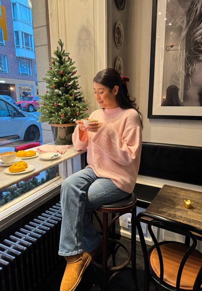 Una suave sudadera rosa y jeans azules son usados por una persona sentada en un taburete de madera oscura. Una mesa presenta café, pasteles y una pequeña decoración dorada.