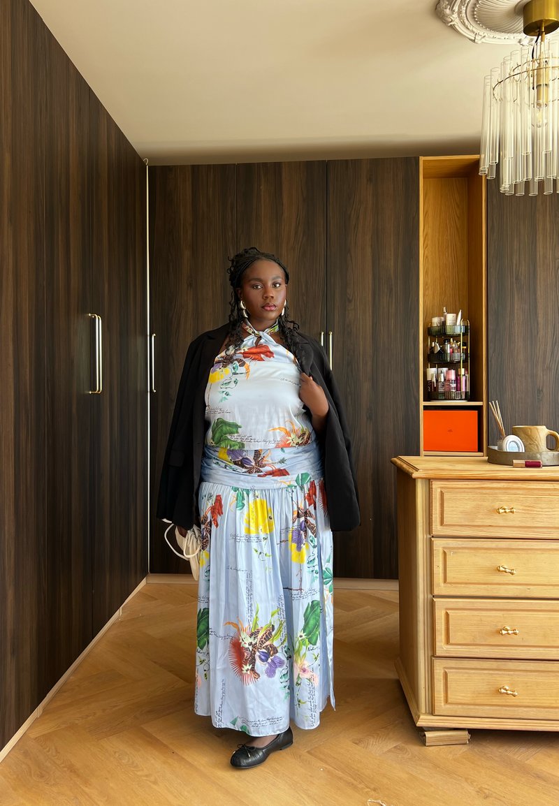 Femme portant une robe fleurie et une veste noire, debout dans une pièce avec des armoires en bois et une commode en bois clair avec des produits de beauté dessus.