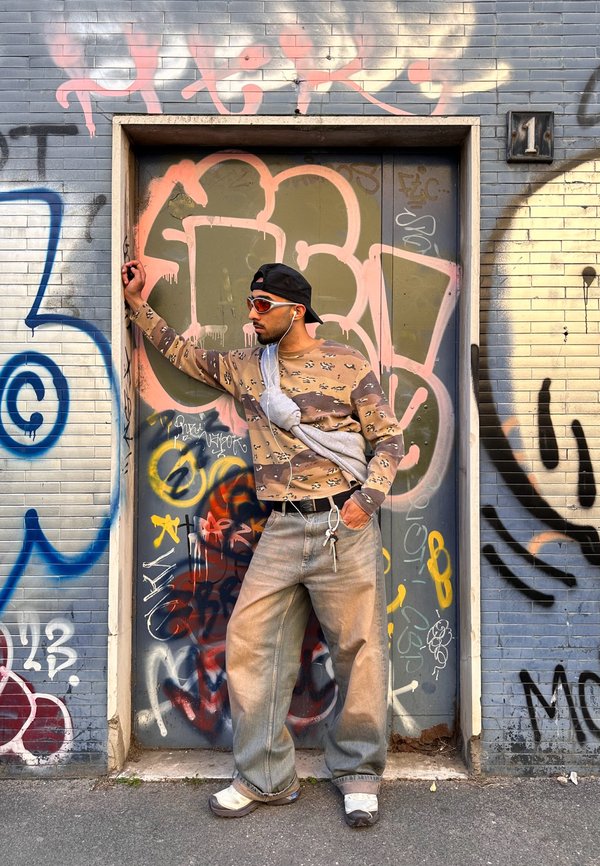 Jeune homme avec des lunettes de soleil et une casquette s'appuie contre une porte couverte de graffitis, portant une chemise à manches longues à motifs, un sac, un jean ample et des écouteurs.