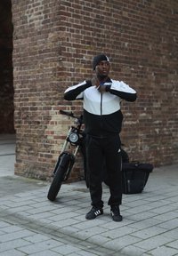 Juodos ir baltos spalvos zip-up striukė su ilgomis rankovėmis, juodos kelnės ir juodi Adidas batai. Šalia asmens pristatomas dviratis.