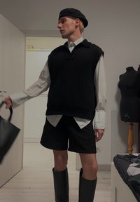 Sweater preto sem mangas sobre uma camisa de manga longa branca, calções pretos e botas de borracha pretas até ao joelho, segurando uma bolsa preta.