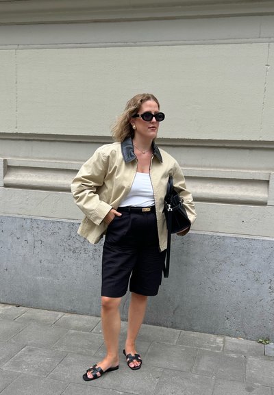 Chaqueta beige con un cuello oscuro, blusa blanca, pantalones cortos negros y sandalias negras. Sosteniendo un bolso negro estructurado. Entorno urbano.