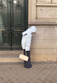 Personne portant une veste à capuche bleu clair et un pantalon rayé, se tenant penchée en avant, tenant un sac à main beige, à côté d'une porte vert foncé et d'un mur en pierre.