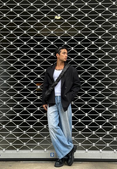 Blazer negro sobre una camiseta blanca sin mangas, jeans anchos de color azul claro y botas negras gruesas, contra un fondo de rejilla metálica.