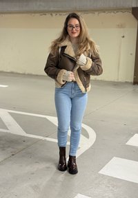 Brun jakke foret med saueskinn med grå mansjetter, beige genser, blå skinny jeans og mørkebrune platåsko i et parkeringshus.