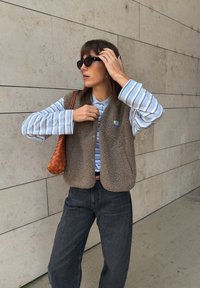 Brūns flīsa veste, svītrota zila un balta krekla ar garām piedurknēm, tumši džinsi un brūna austā soma. Fons veido beidžs sienu paneļi.