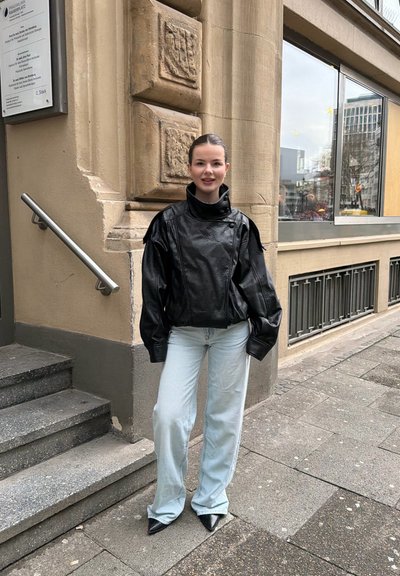 Una joven está de pie en una acera de la ciudad, llevando una chaqueta de cuero negra, pantalones vaqueros anchos de lavado claro y zapatos negros puntiagudos cerca de unos escalones de piedra.