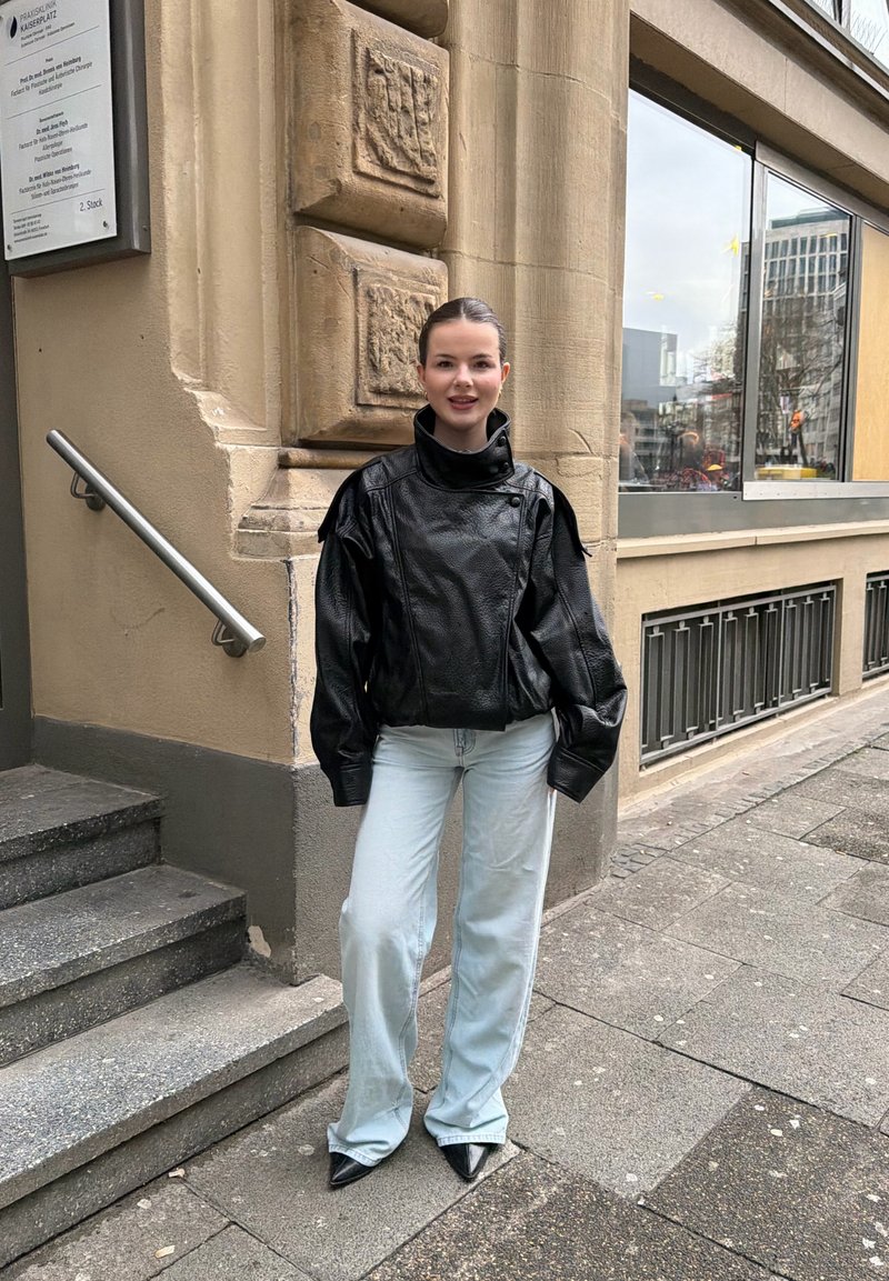 Jeune femme debout sur un trottoir en ville, portant une veste en cuir noire, un jean large bleu clair et des chaussures noires à bouts pointus près de marches en pierre.