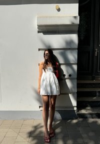 Robe d'été blanche avec un corsage ajusté et une jupe ample, accompagnée de sandales rouges à lanières. Lunettes de soleil carrées et un sac à main rouge ; devant un mur blanc.