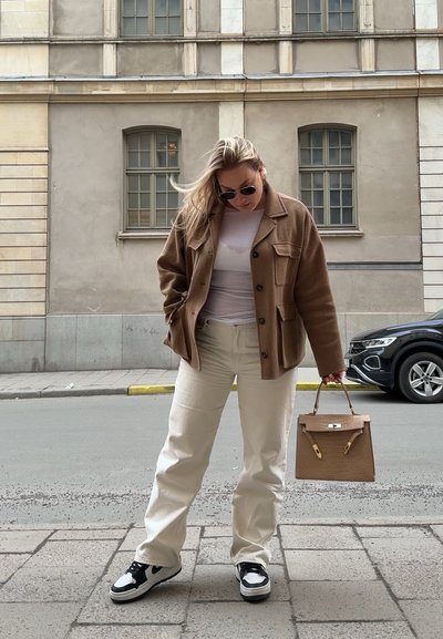 Chaqueta de mezcla de lana marrón sobre una camisa blanca, combinada con pantalones crema y zapatillas en blanco y negro. Sosteniendo un bolso de textura marrón.