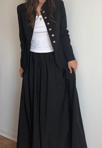 Schwarzer Blazer mit goldenen Knöpfen, getragen über einem weißen Tanktop und einem schwarzen, bodenlangen, plissierten Rock. Glatte Textur mit taillierter Passform.