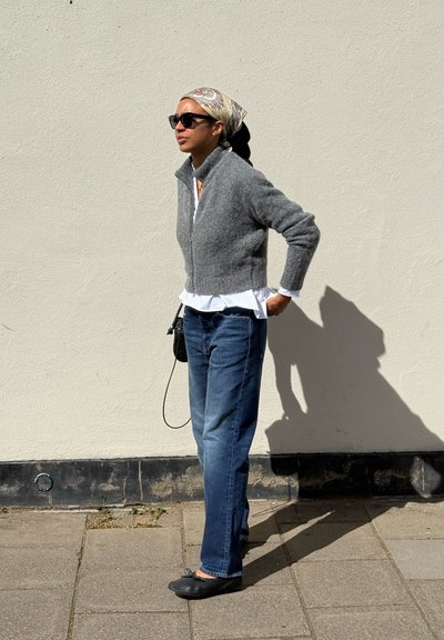 Mujer con gafas de sol, pañuelo en la cabeza, suéter gris con cremallera, camisa blanca, vaqueros azules y zapatos planos negros, de pie en una acera frente a una pared beige.