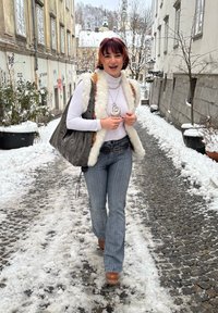 Mannequin portant un haut blanc à manches longues, un gilet en fourrure et un jean évasé à rayures, tenant un sac gris à motifs. Chemin pavé couvert de neige.