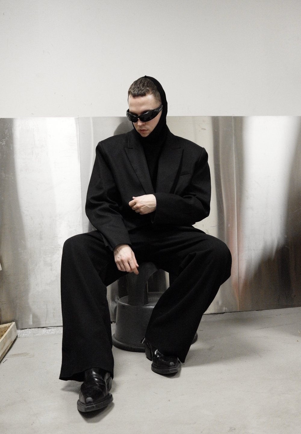 Veste de costume noire surdimensionnée et haut à col montant, pantalon noir à coupe décontractée, chaussures noires brillantes et lunettes de soleil foncées sur un fond métallique.
