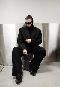 Veste de costume noire surdimensionnée et haut à col montant, pantalon noir à coupe décontractée, chaussures noires brillantes et lunettes de soleil foncées sur un fond métallique.