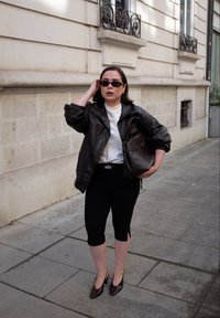 Vrouw in een zwart leren jasje, zwarte capri-broek en hakken, met een grote bruine tas op een stoep in de stad met een stenen gebouwen op de achtergrond.