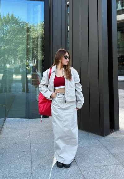 Conjunto de chaqueta y falda de rayas en gris claro, top rojo corto, zapatos negros, mochila grande roja, gafas de sol, de pie sobre una superficie pavimentada.