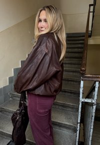 Barna bőrdzseki túlméretezett ujjal, bordó nadrággal párosítva. A háttérben lépcső és fakorlát látható.