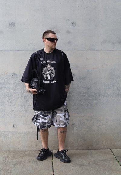 Camiseta negra de gran tamaño con impresiones gráficas en blanco, pantalones cortos de camuflaje, zapatillas negras y una bolsa negra. Superficie lisa y fondo neutro.