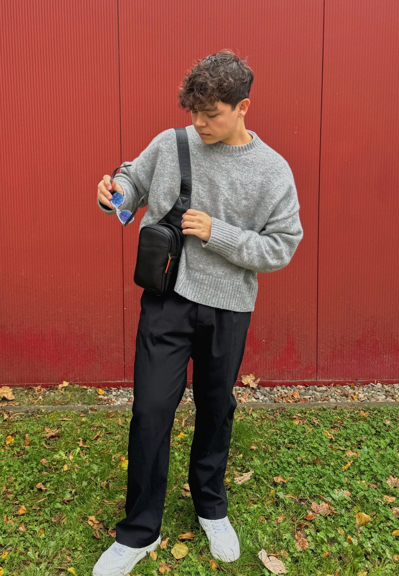 Γκρι πλεκτό πουλόβερ, μαύρη τσάντα ώμου, μαύρα παντελόνια, κρατώντας γυαλιά ηλίου. Απλός σχεδιασμός, χαλαρή εφαρμογή, υφασμάτινο υλικό. Φόντο με πράσινο γρασίδι.