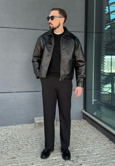 Chaqueta de cuero negra sobre un suéter de punto negro, combinada con pantalones negros y zapatos negros brillantes. El entorno presenta una pared gris y cristal.