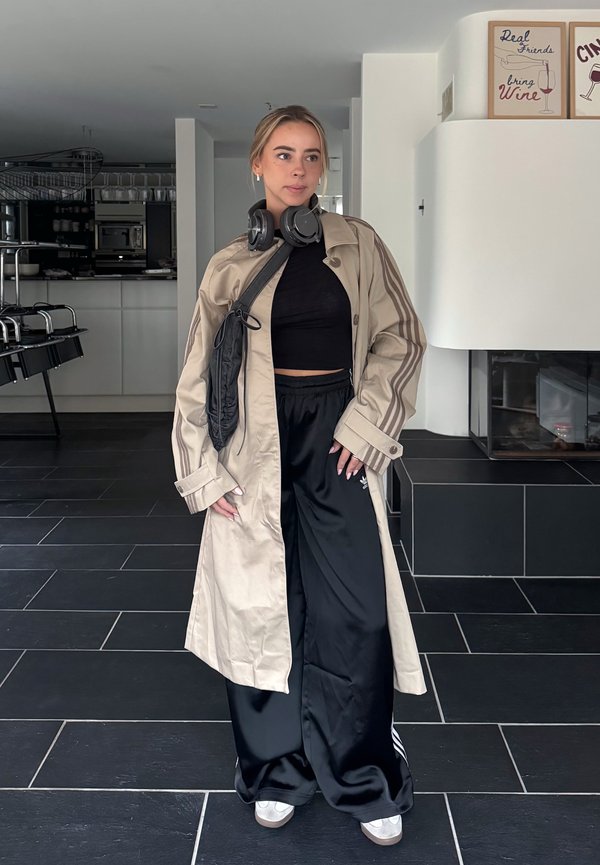 Joven mujer con un abrigo largo beige, top corto negro y pantalones anchos negros está en el interior con auriculares alrededor del cuello y una pequeña bolsa negra sobre el hombro.