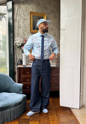Camisa abotonada azul claro con detalles de costura roja, jeans de pierna ancha azul oscuro, gorra azul claro y zapatillas deportivas. Entorno interior con decoración.