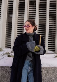 Γυναίκα με μοβ γυαλιά, σκουρόχρωμο παλτό, γκρι πουλόβερ, πράσινα γάντια και τζιν, στέκεται έξω κοντά σε μοντέρνο κτίριο με χιόνι και φυτά.