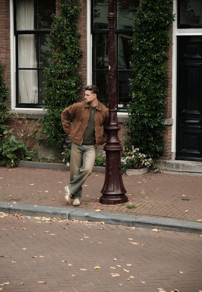 Chaqueta marrón sobre un suéter verde, jeans claros desgastados y zapatillas beige. De pie junto a una farola color marrón con vegetación de fondo.