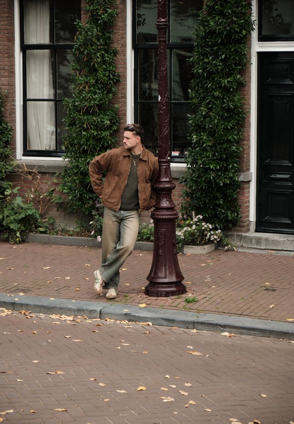 Brun jakke over en grøn sweater, lyseblå slidte jeans og beige sneakers. Står ved en maroon gadelampe med grønne planter bag sig.