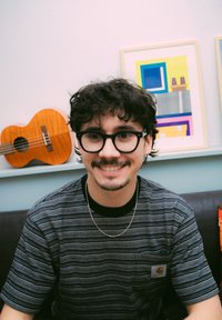 Jovem com cabelo escuro e encaracolado, óculos, camisa às riscas e colar de corrente prateada, sorrindo em ambientes fechados com um ukulele e arte abstrata ao fundo.
