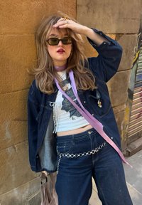 Τζάκετ από τζιν με γκρι εσωτερικό, λευκή μπλούζα με σχέδιο, φαρδύ παντελόνι τζιν και ασημένια ζώνη με αλυσίδα. Λεβάντα κορδέλα ως διακόσμηση.